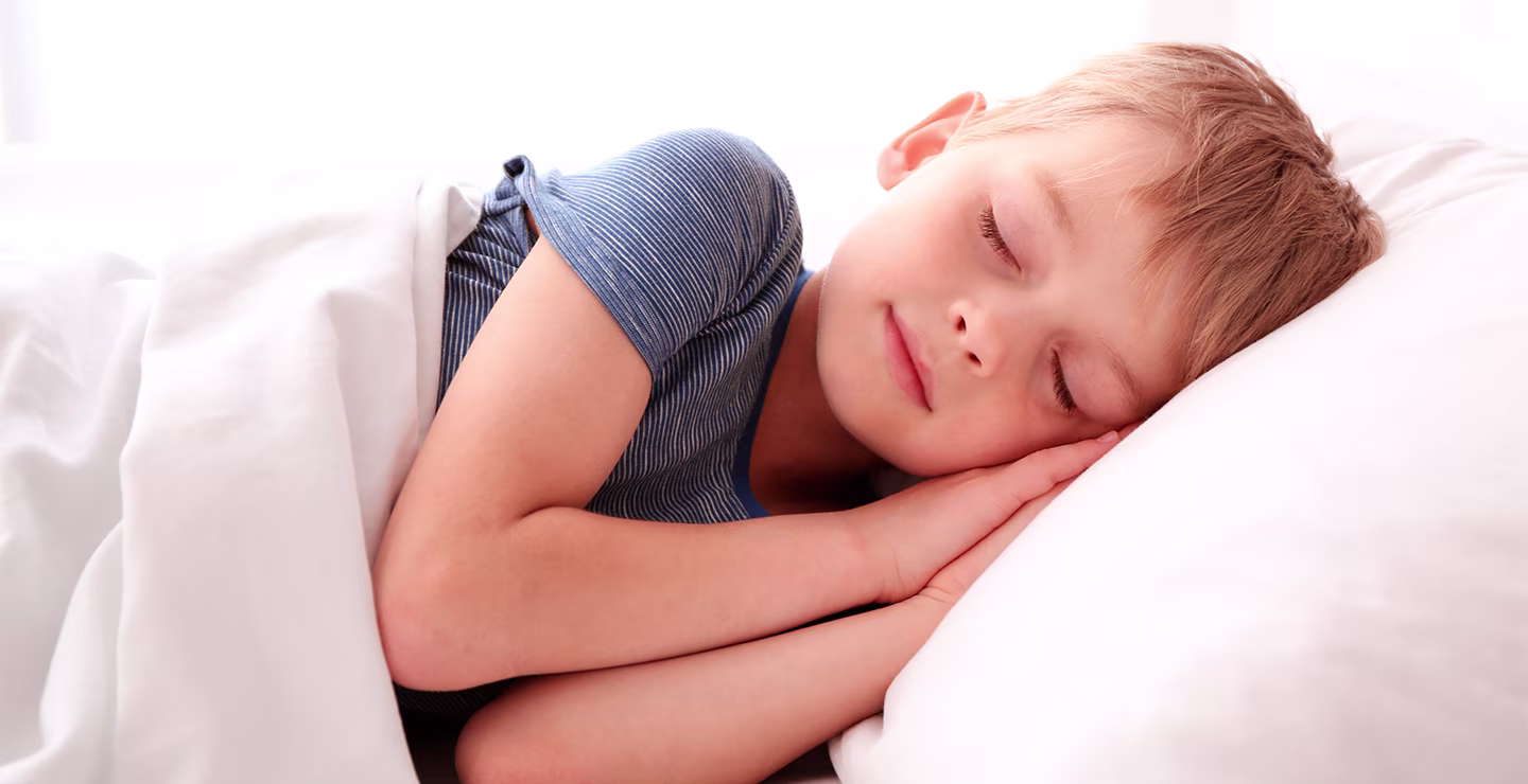 A boy lying on a bed with white bedding wearing a blue striped shirt in a bright bedroom.