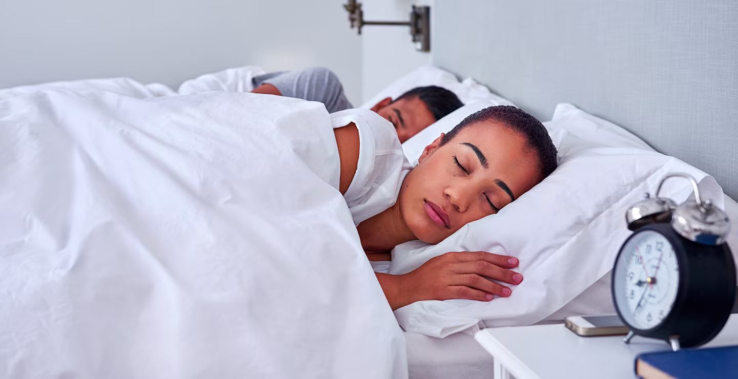 Two people sleeping in a bed with white bedding and a nightstand with clock and book.