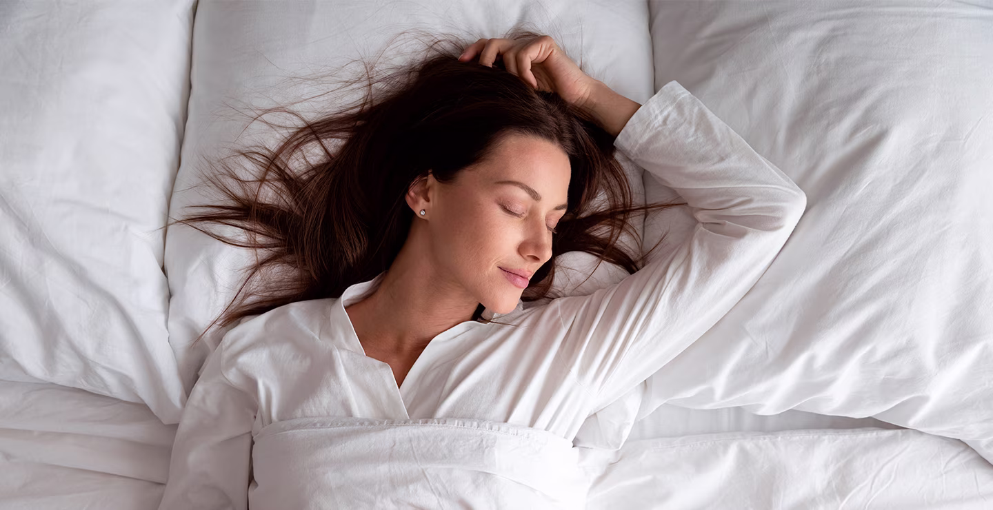 A woman with brown hair sleeps peacefully in a white bed, her hand resting near her head.