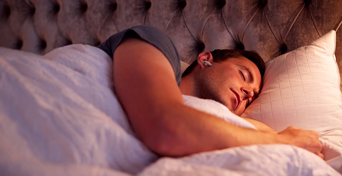 A man is sleeping peacefully in a bed and a tufted headboard while wearing a wireless earphone.