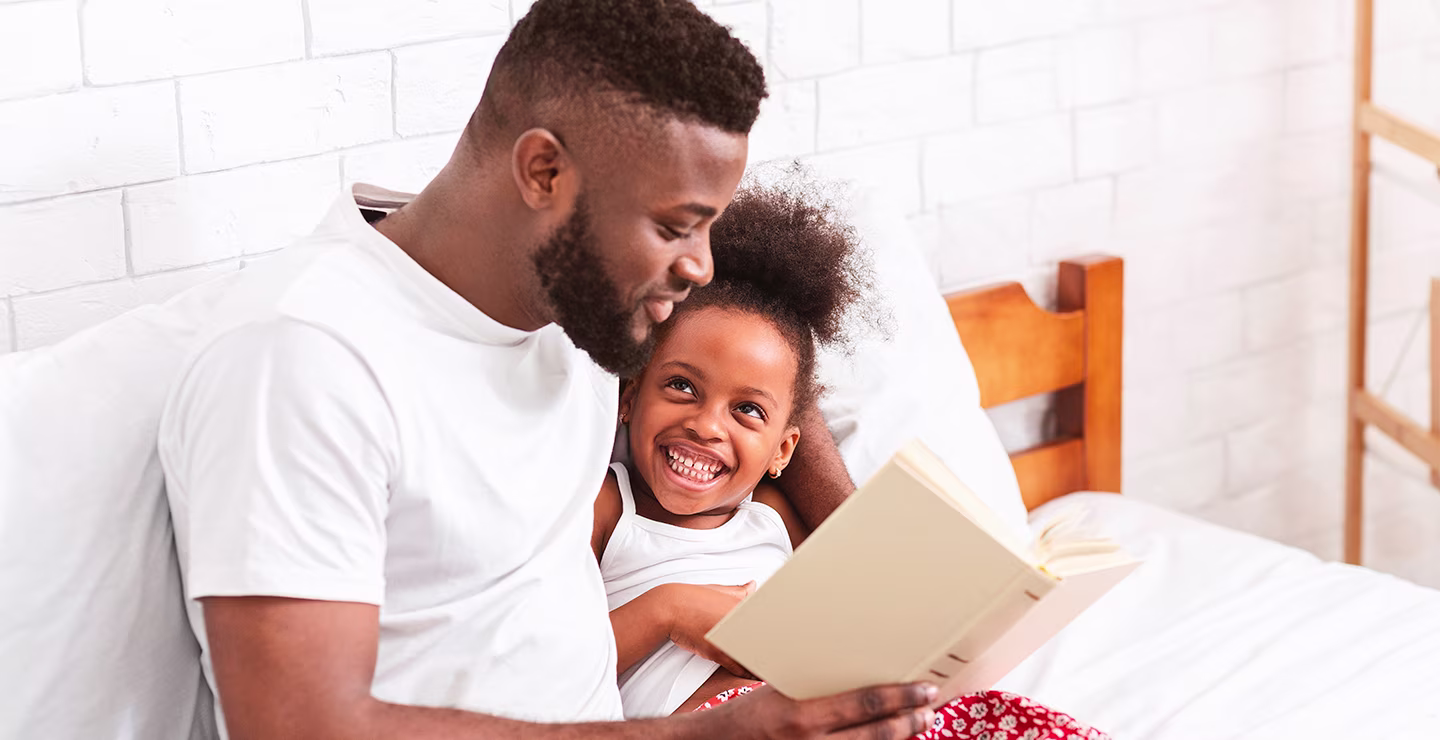African father reading with his adorable little daughter at home in bed.